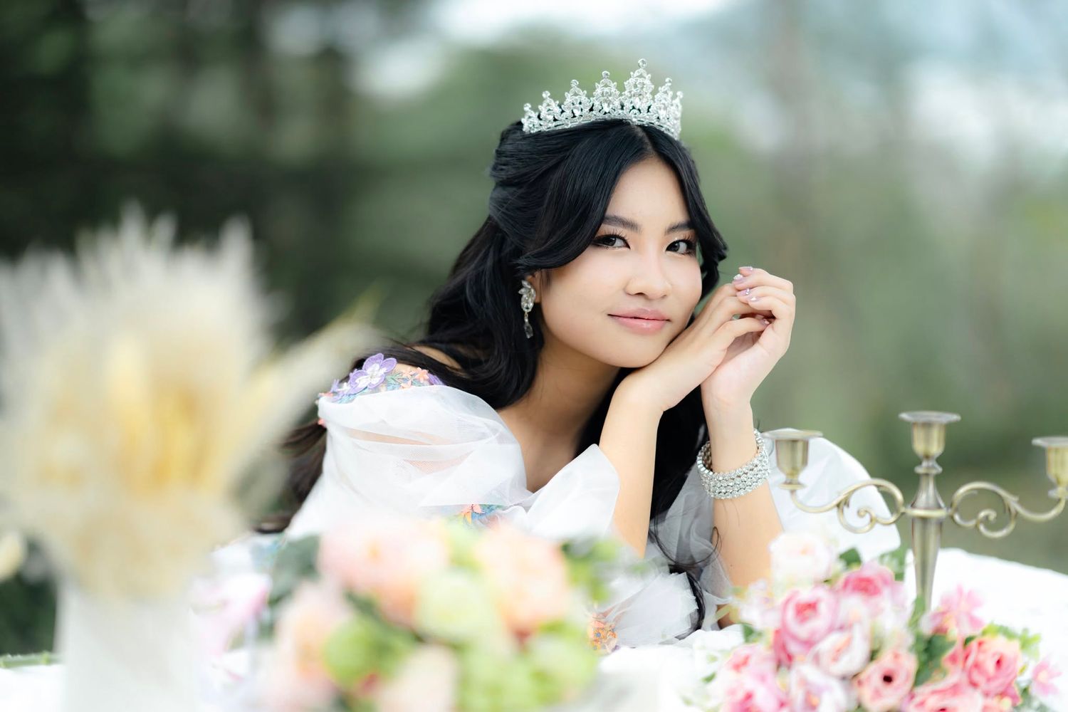 Young Asian woman wearing a crystal tiara and white dress, posing gracefully at a floral table outdoors.