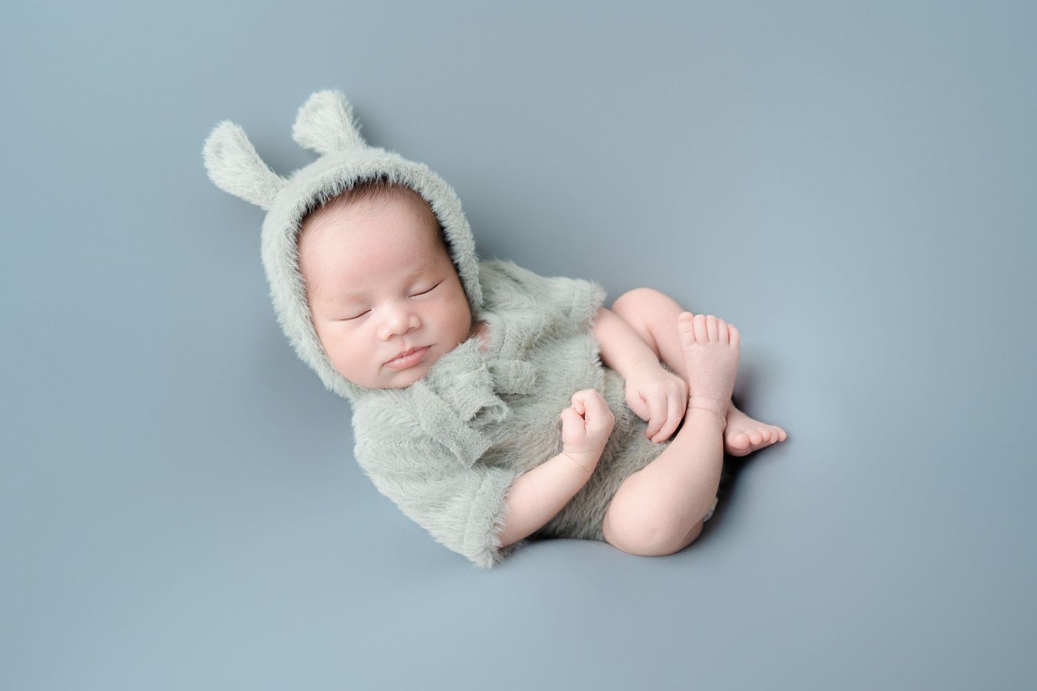 Infant in light gray outfit sleeping on soft fabric with feet visible.
