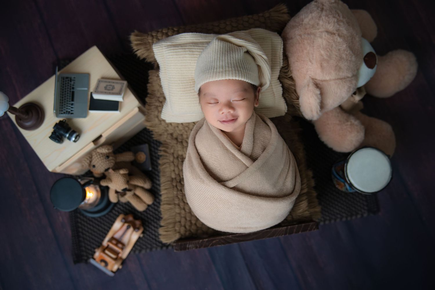 Newborn photography setup with beige swaddle, vintage toys and soft lighting on dark backdrop.