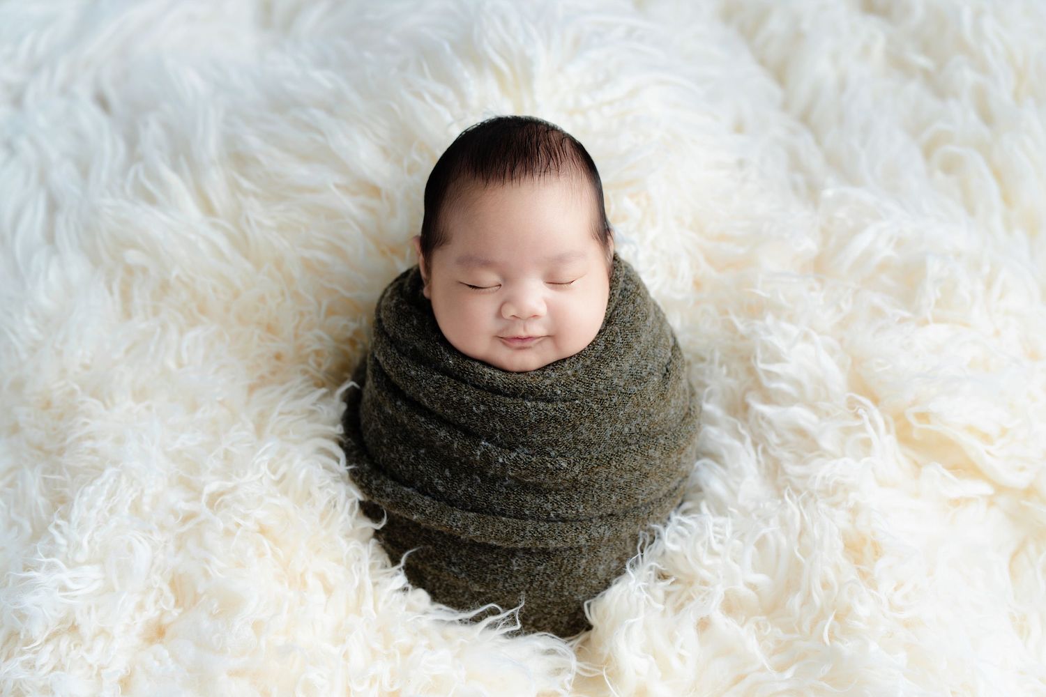 A peaceful baby swaddled in olive green fabric rests on soft white fur during a newborn photo session.