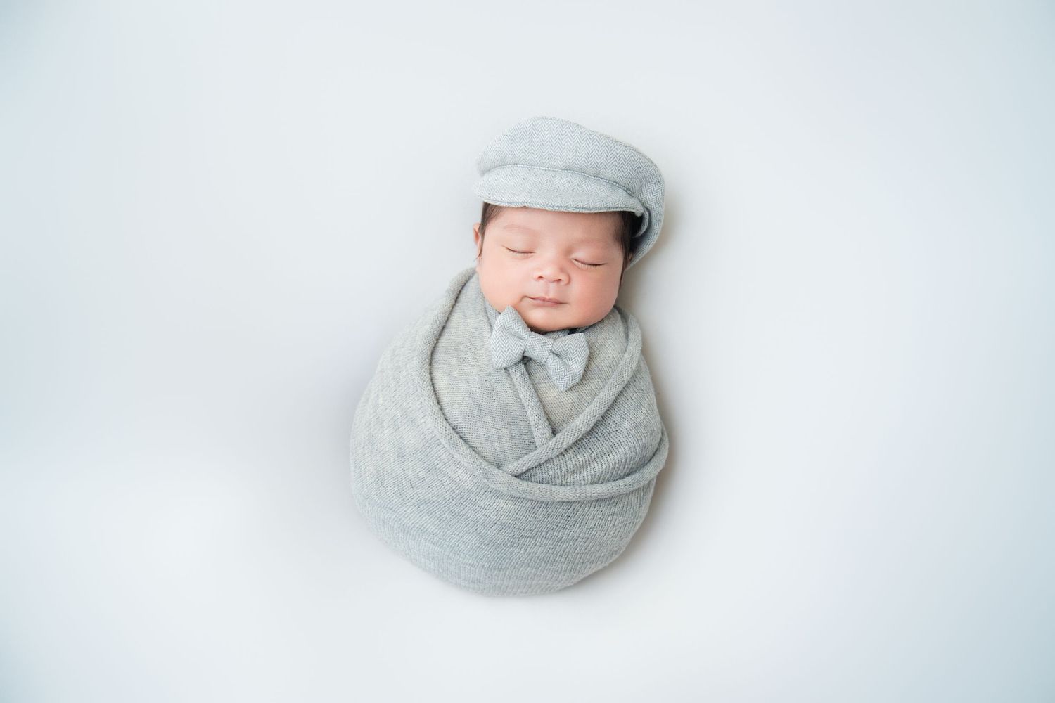 Sleeping newborn wrapped in gray fabric with matching cap on white background.