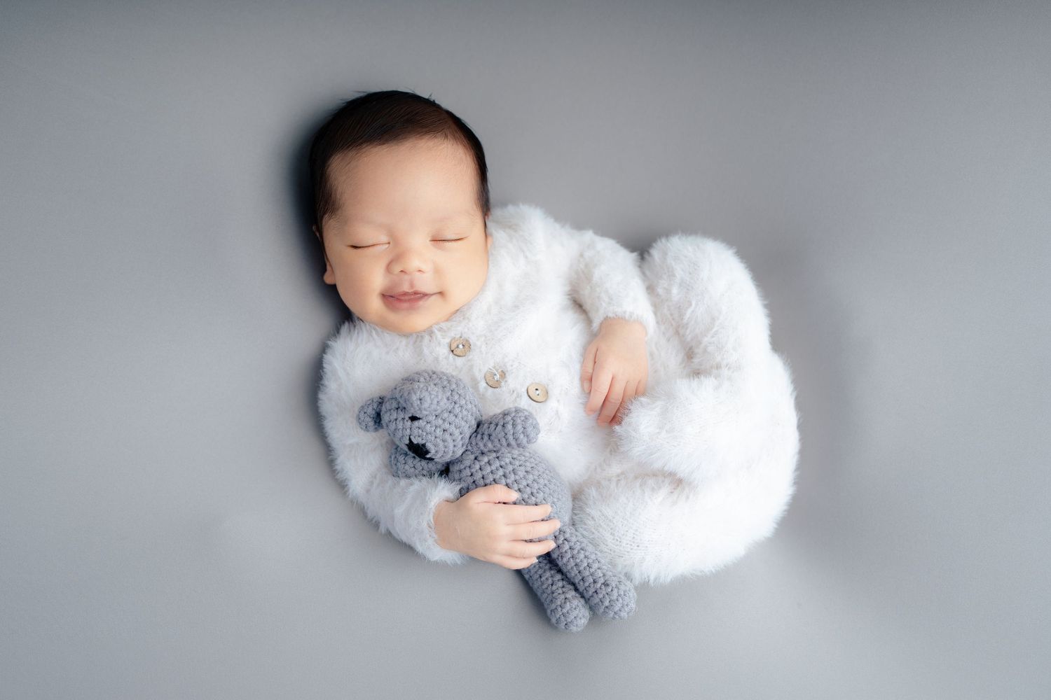 Newborn baby wearing white outfit sleeping peacefully on soft gray backdrop in professional studio setting.