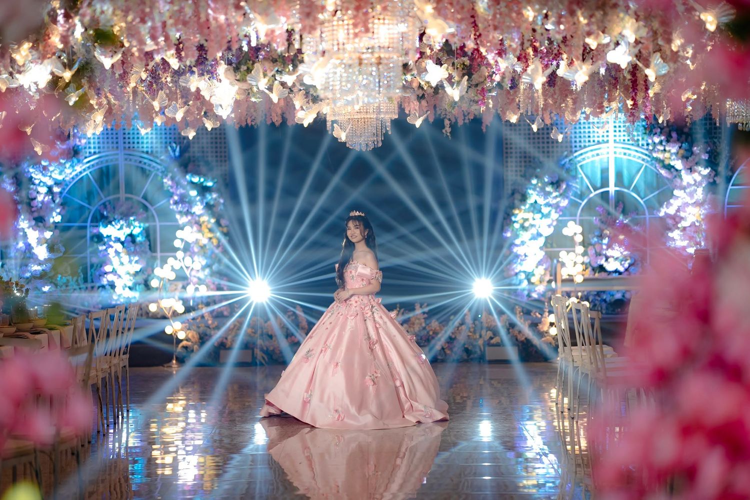 Woman in pink ball gown stands on reflective floor surrounded by floral arch and dramatic stage lighting at elegant event.