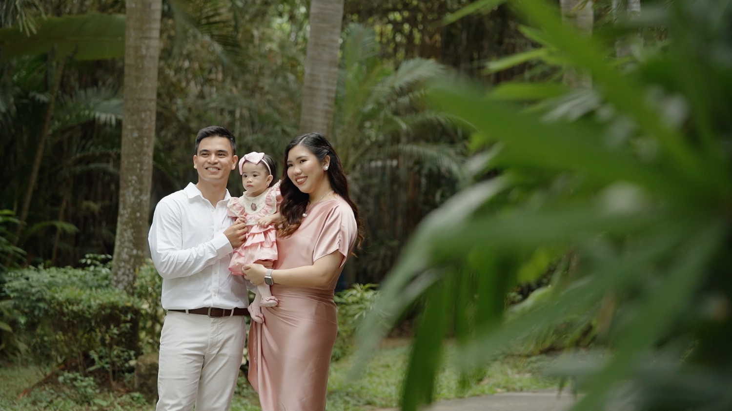 Happy Asian family with parents holding baby girl posing outdoors in lush green tropical garden setting.