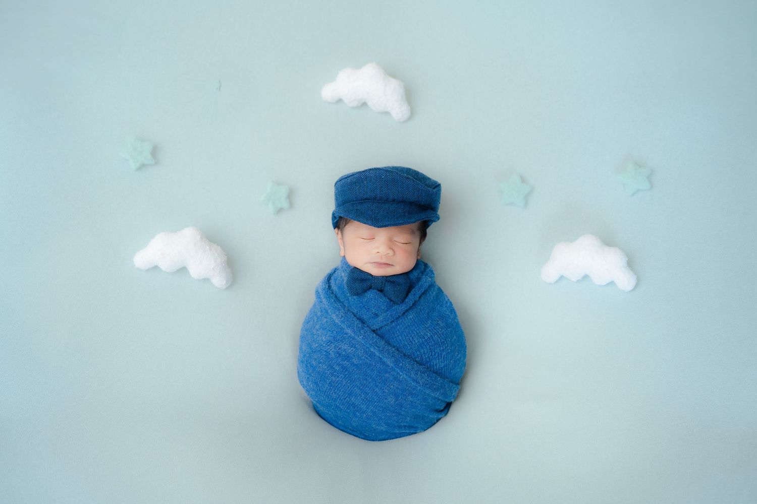 Newborn photography with baby in blue wrap and cap surrounded by white cloud decorations.