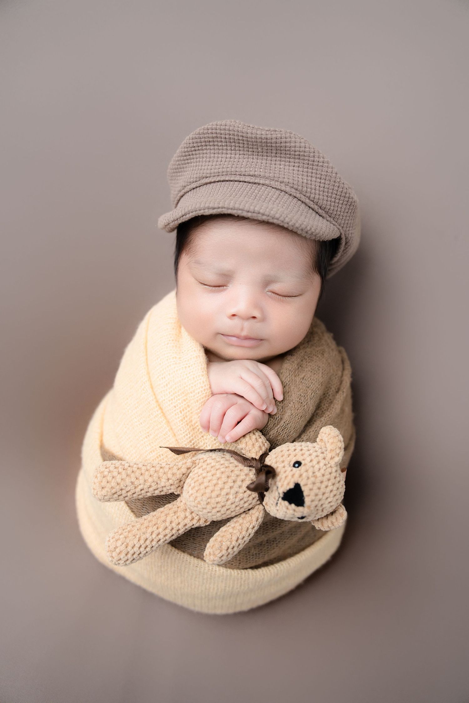 Sleeping infant wearing beige cap with knitted teddy bear on neutral background.