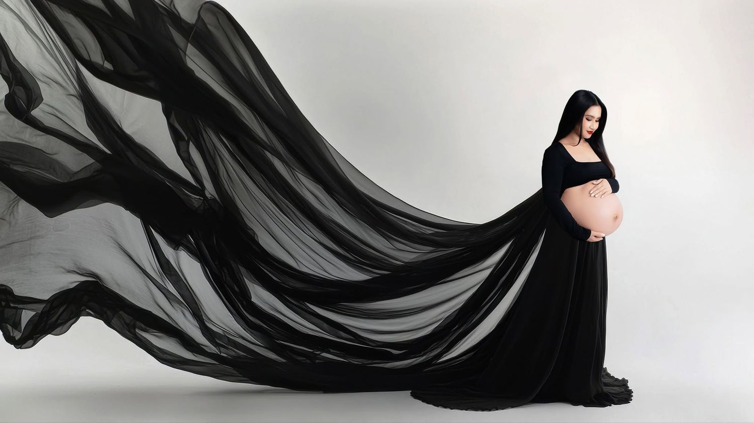 Pregnant woman in flowing black maternity gown with dramatic fabric billowing against white background.