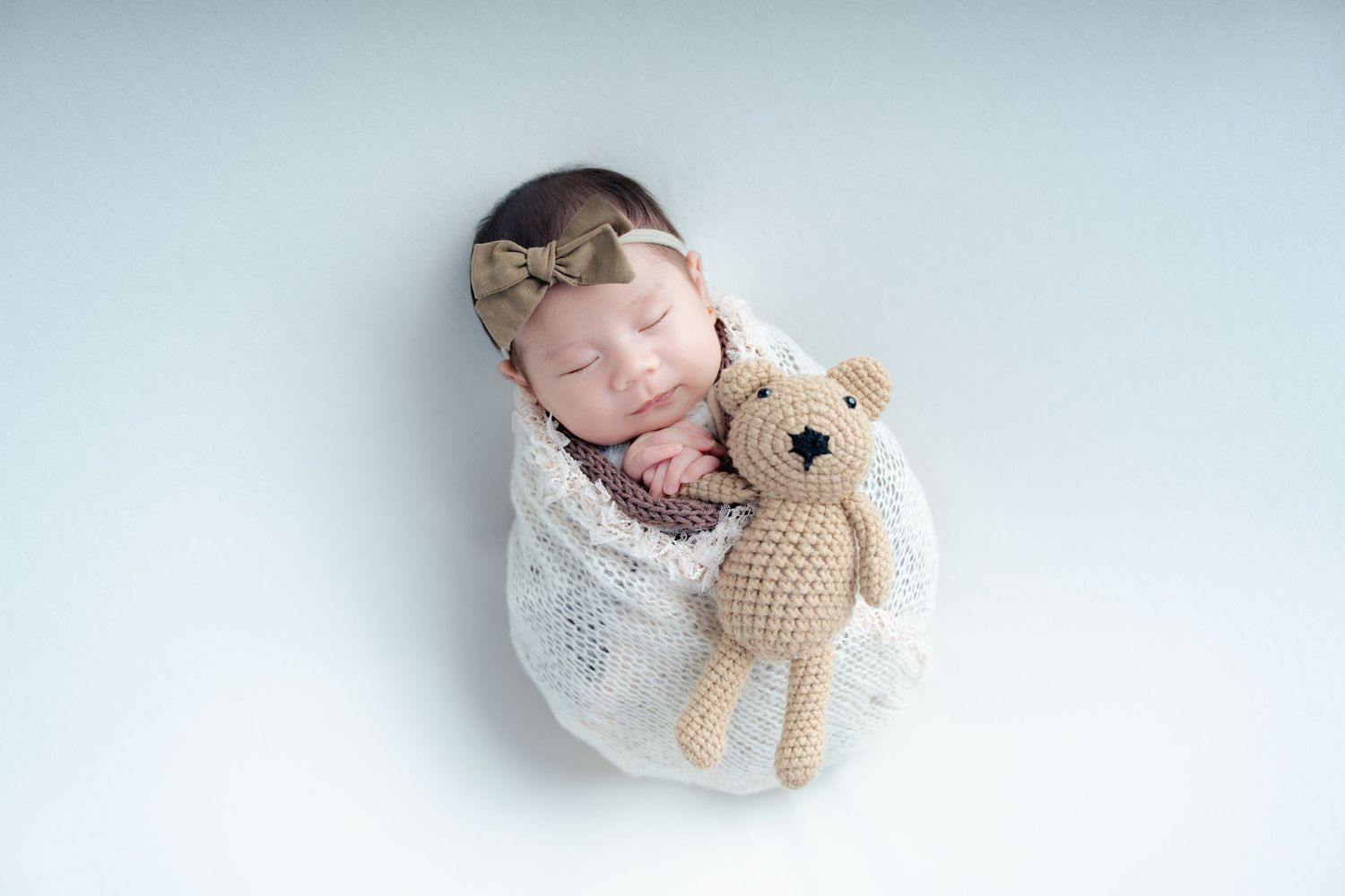 Newborn baby wrapped in white blanket sleeping peacefully with knitted teddy bear on light background.