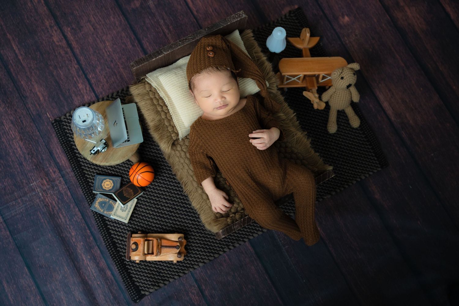 Brown-clothed baby resting on blanket surrounded by wooden toys and vintage accessories.
