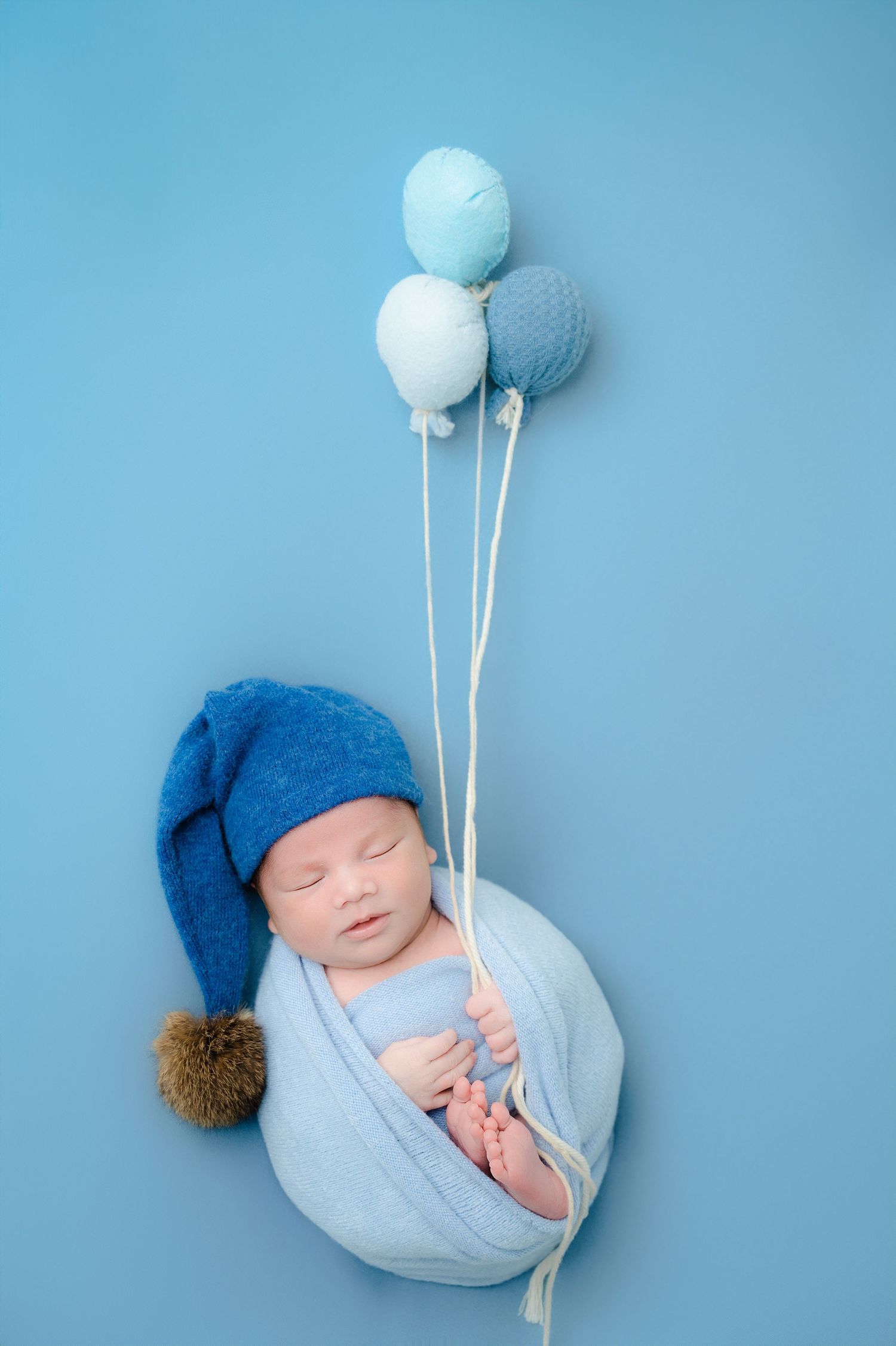 Newborn wearing blue hat with white balloons on blue background.