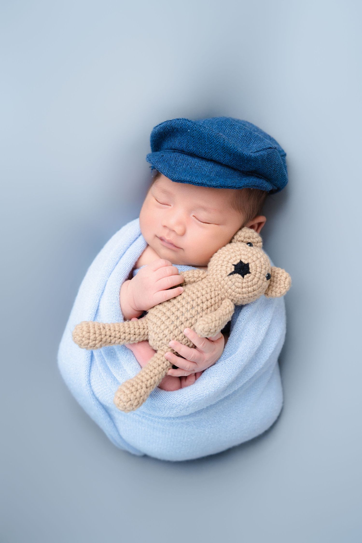 Newborn in blue cap cuddling tan knit teddy bear while wrapped in light blue blanket.