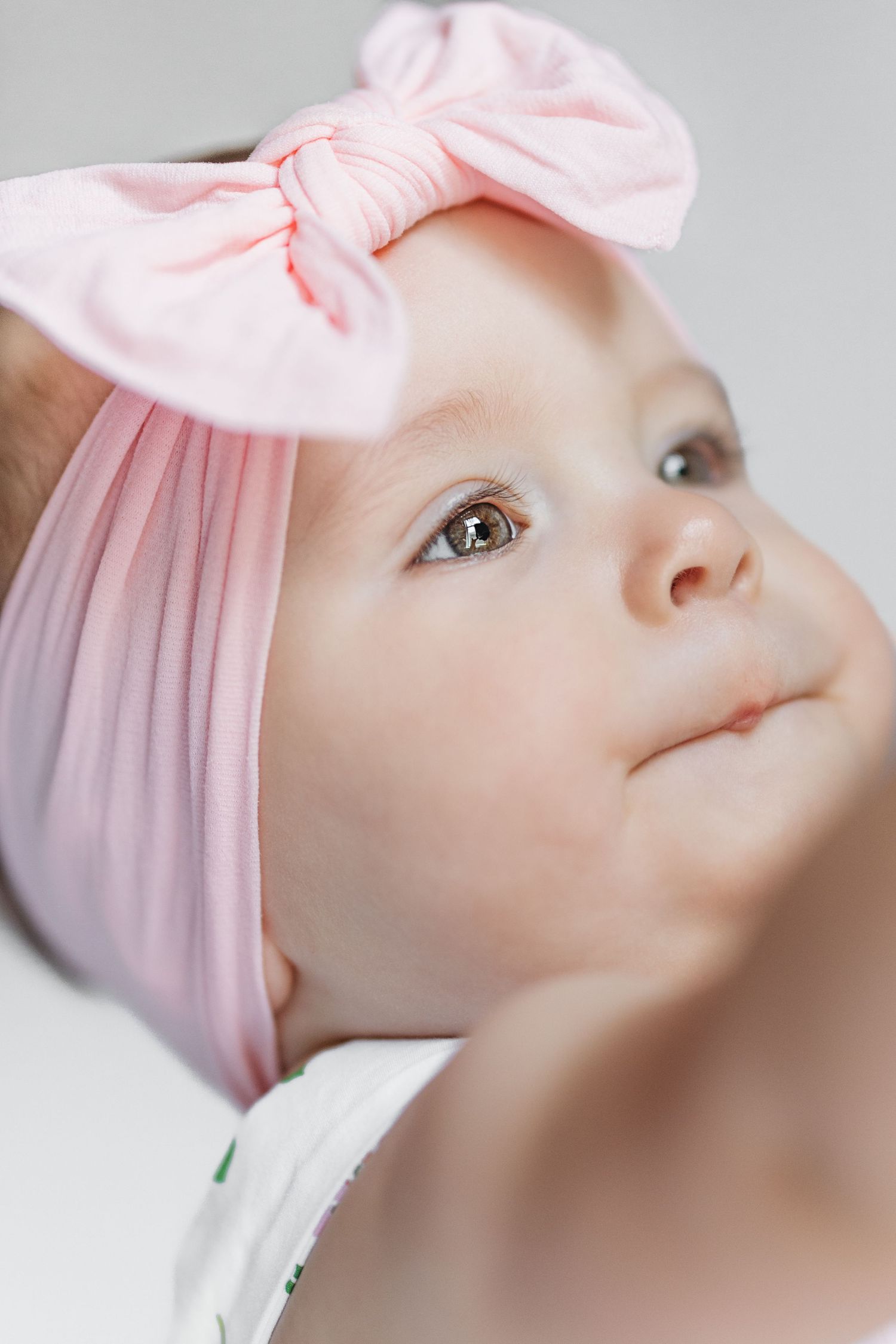 Close-up portrait of an adorable baby girl wearing a pink bow headband, looking up with curious brown eyes.