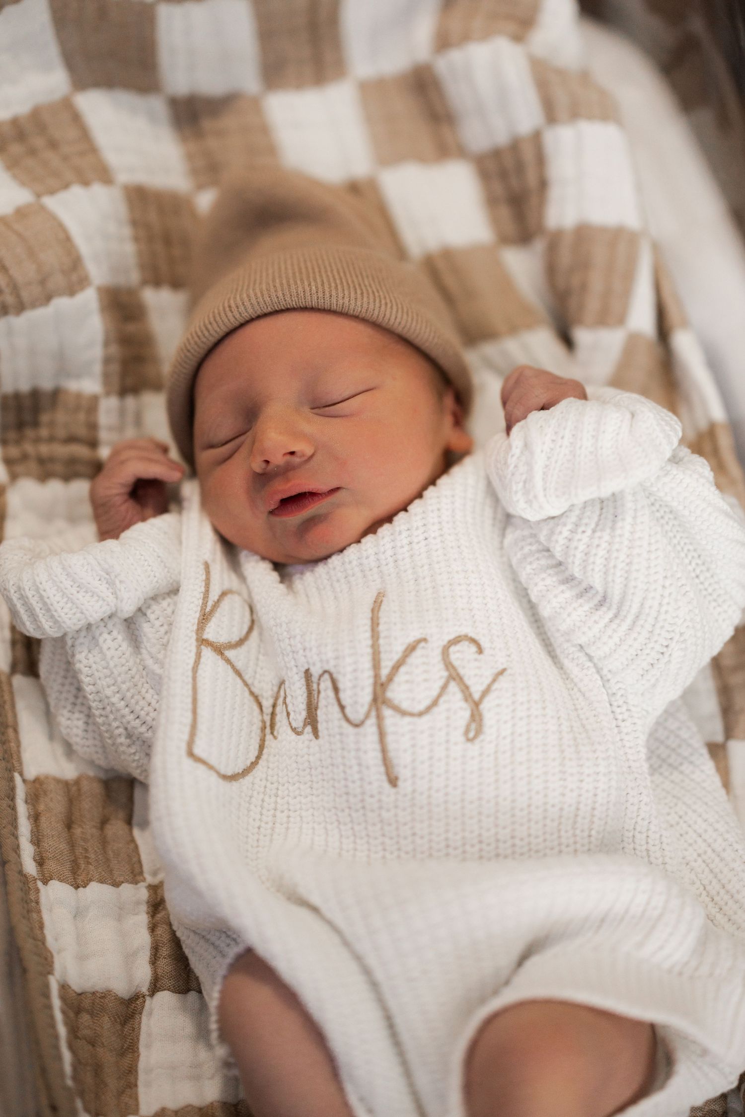 Newborn baby sleeping peacefully in white knit personalized romper with beige hat on checkered blanket.