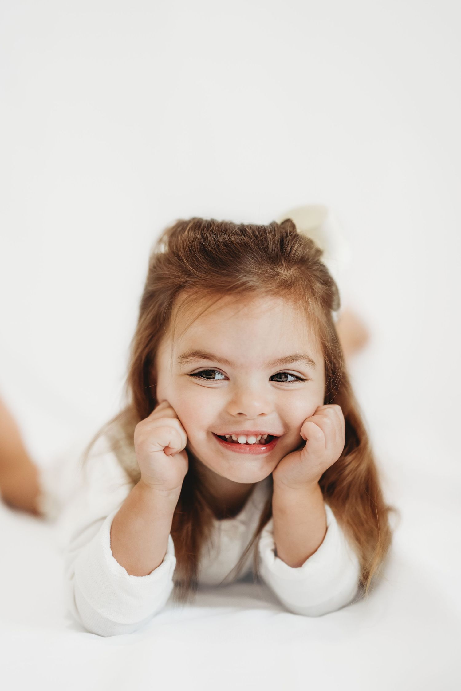 Smiling little girl with long brown hair resting her chin on her fists, lying on a white surface.