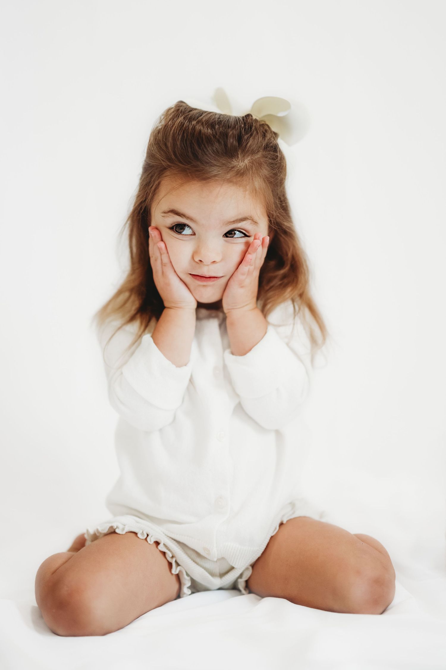 A cute little girl in a white outfit sits cross-legged with hands on cheeks, posing adorably against a white background.