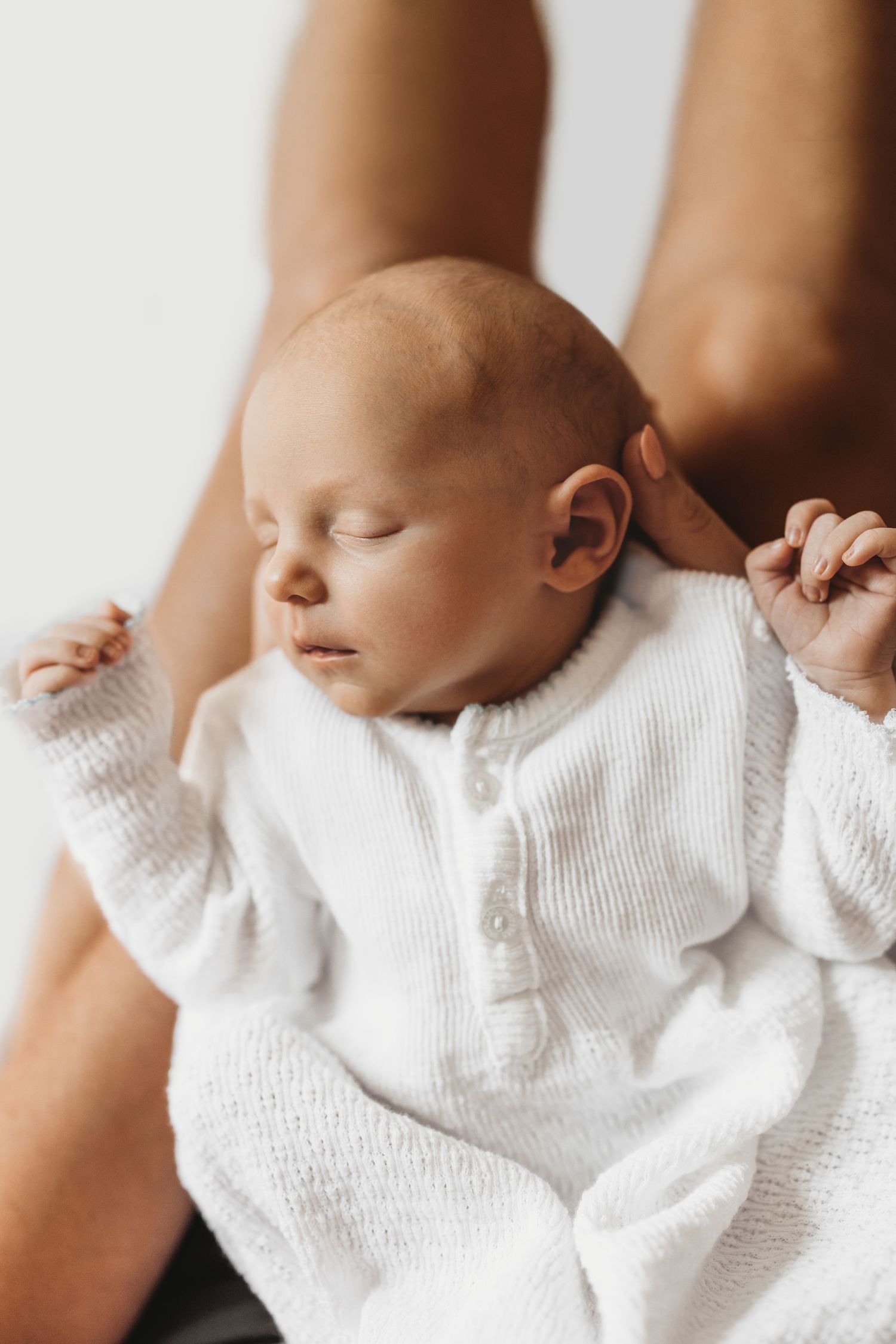 Peaceful newborn baby sleeping in white knit onesie, cradled gently in parent's hands against white background.