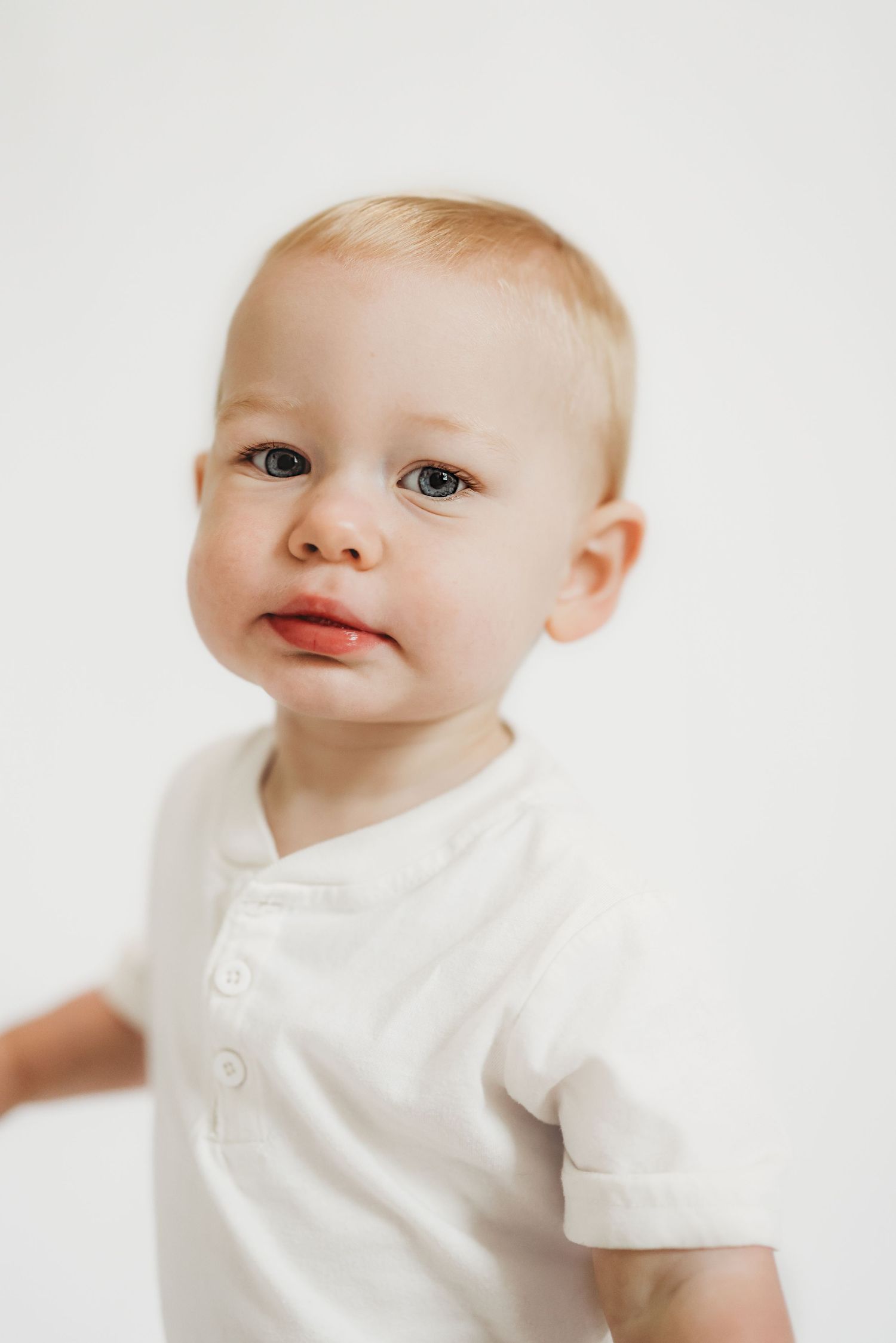 Adorable baby boy with blonde hair and blue eyes wearing a white shirt, posed against a white background.