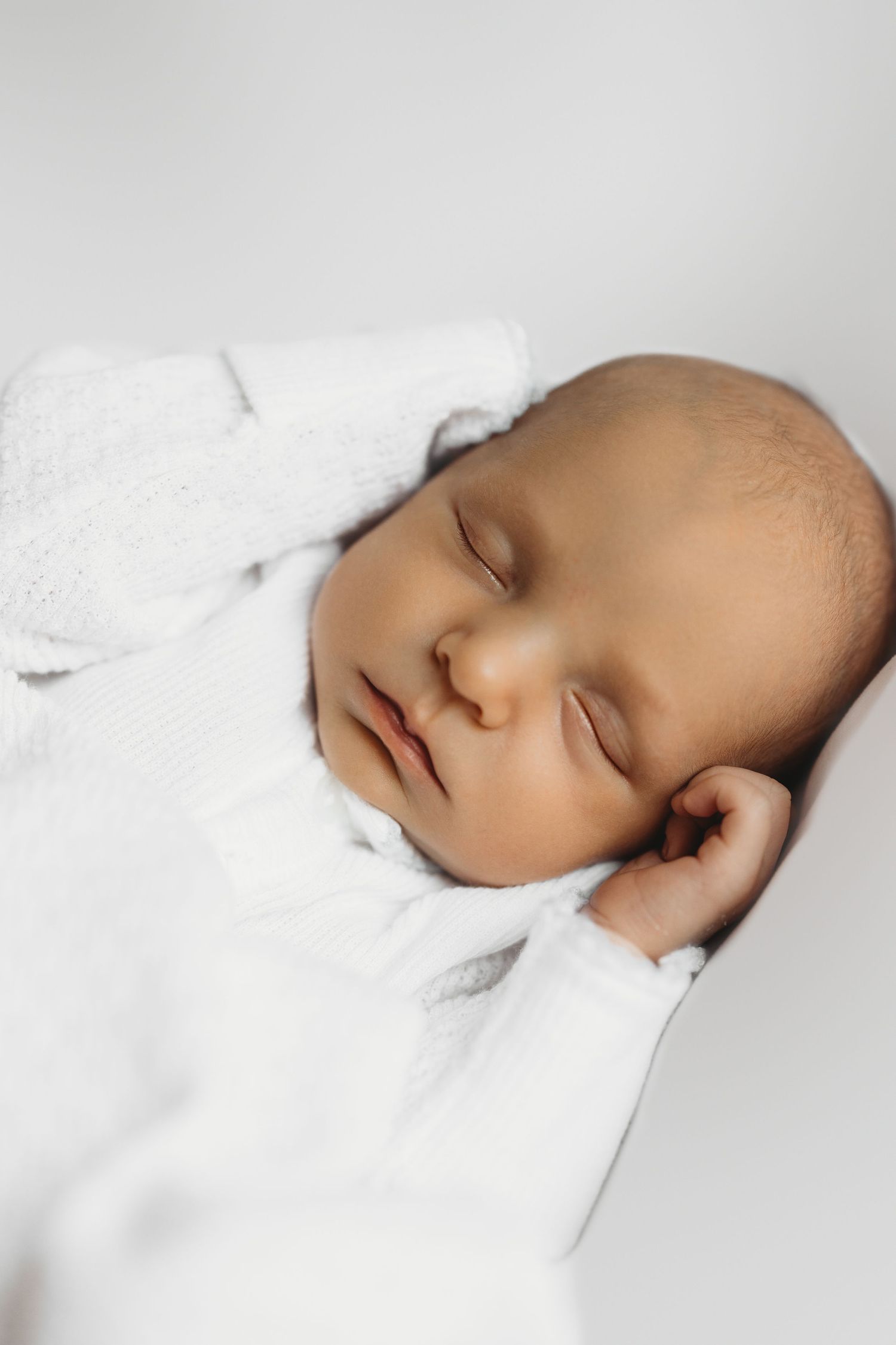 Peaceful newborn baby sleeping in a white onesie against a soft white background.