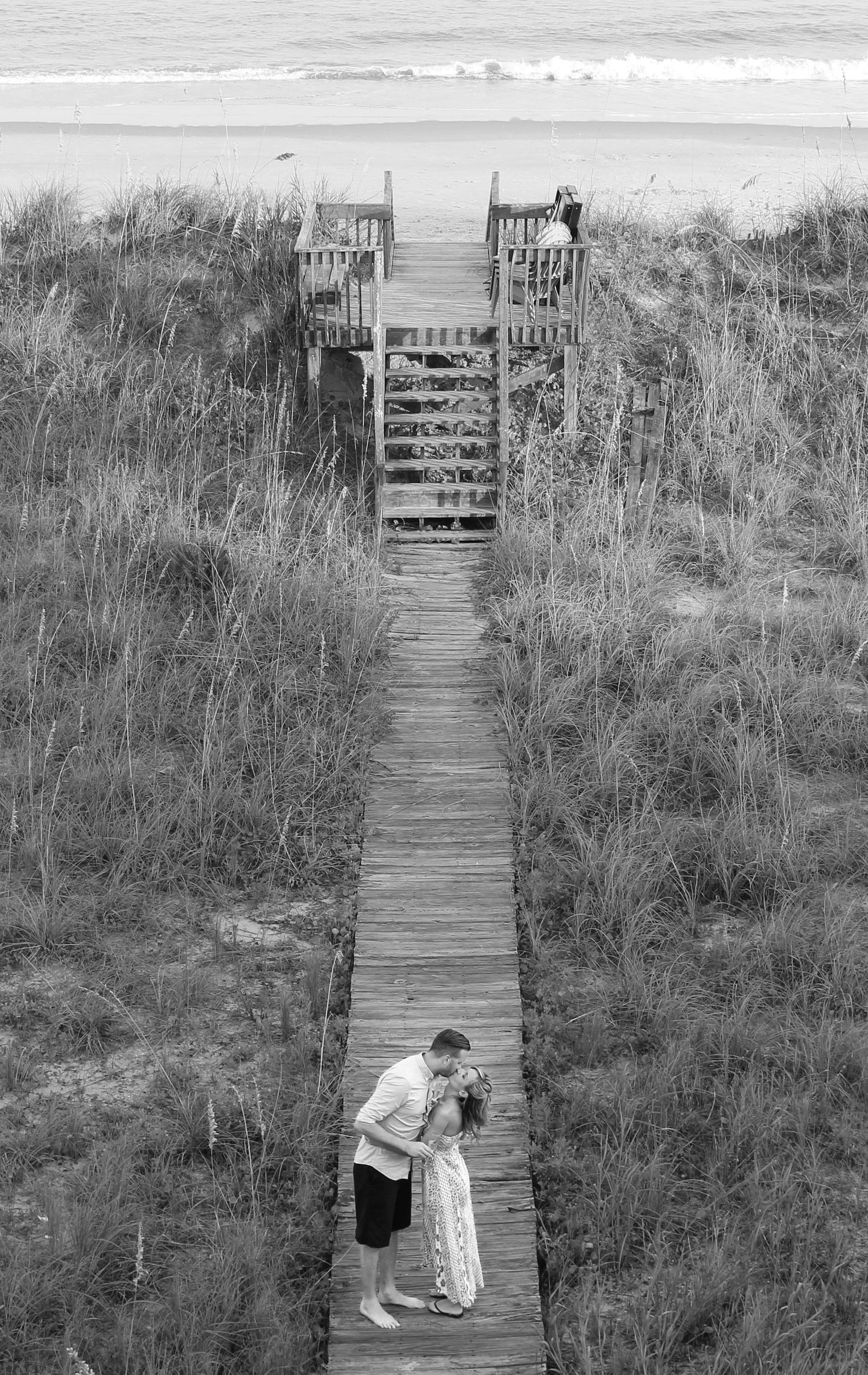 Black and white enagement photograph of a woman and man at a beach on a long dock.