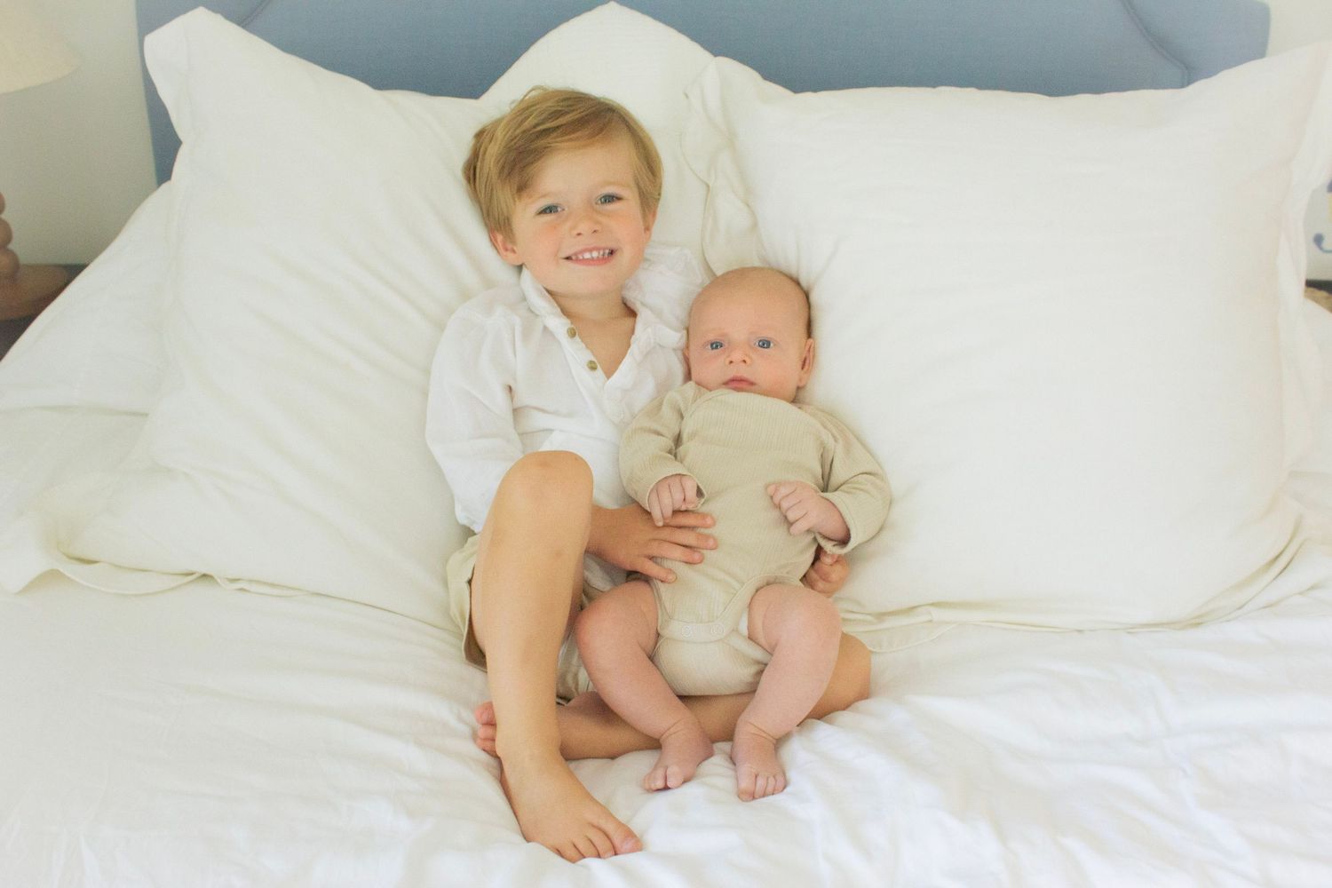 Big brother sitting on a white bed holding his newborn baby sibling during a family photography session in Fairfield County, CT.