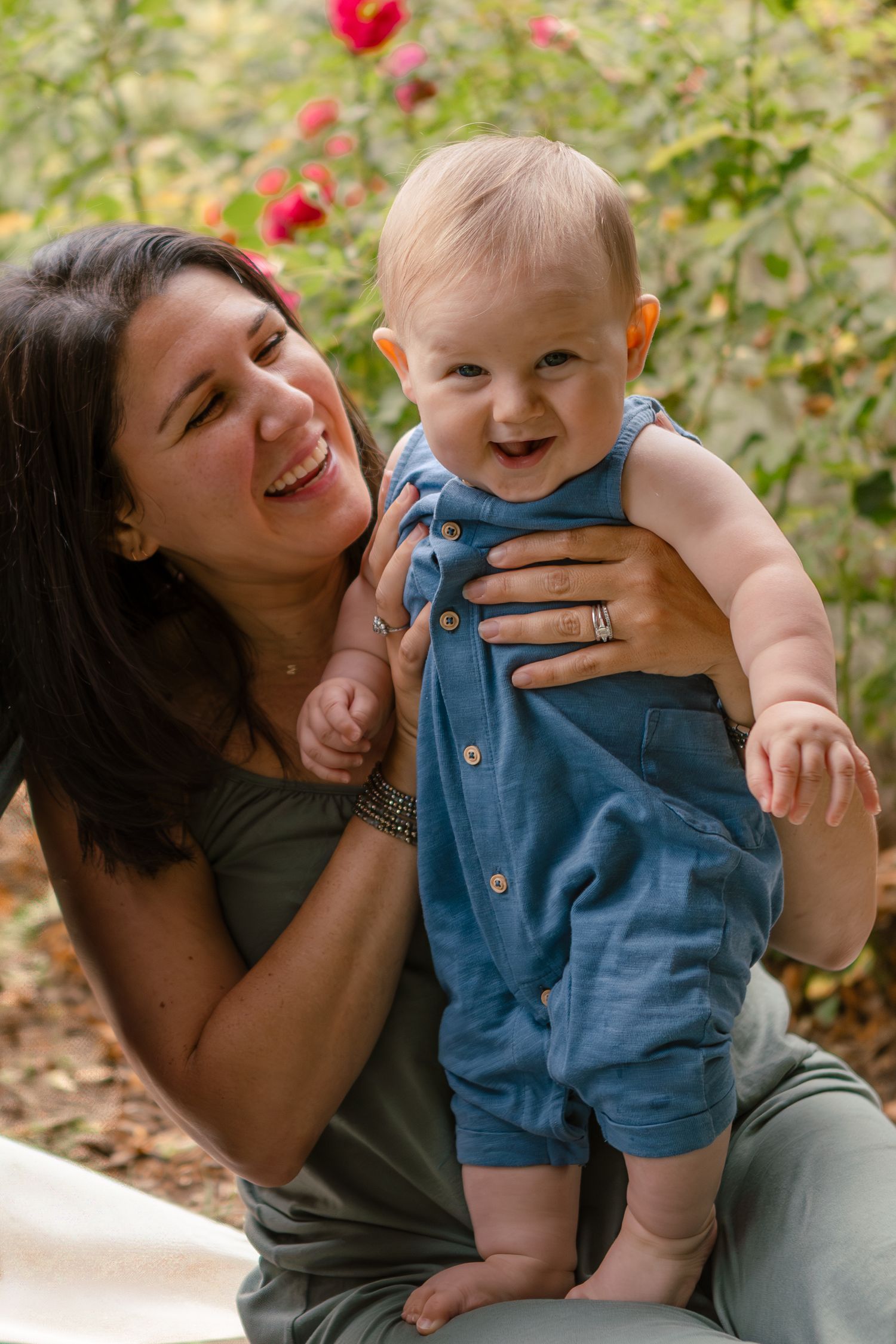 Mother sitting in park and holding baby upright on her lap looking at baby and smiling softly, while baby faces forward giving a cheeky grin