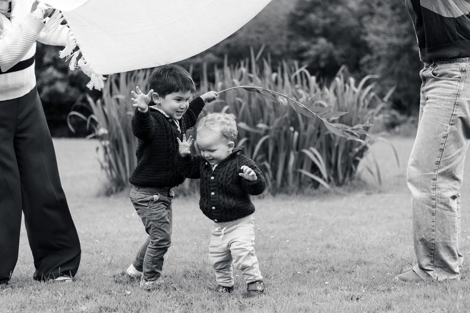 Toddler brother reaches out to grab and hug little brother in park while mothers look on