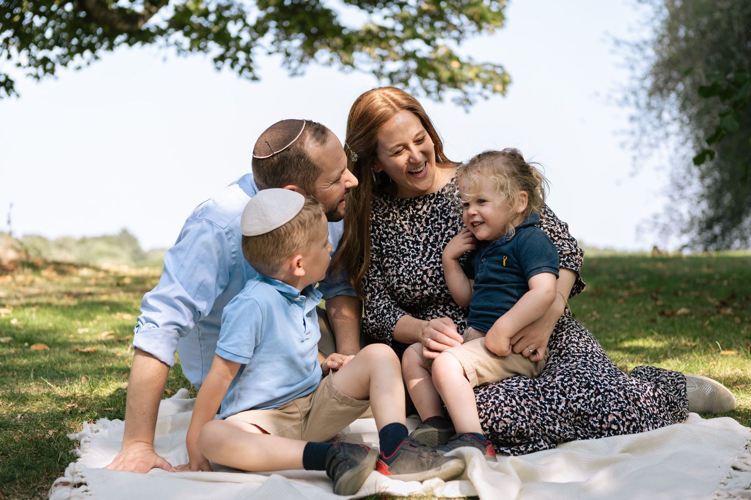 Family of four on blanket in park, with mother, father and older brother smiling at toddler sitting on mother's lap, toddler smiling