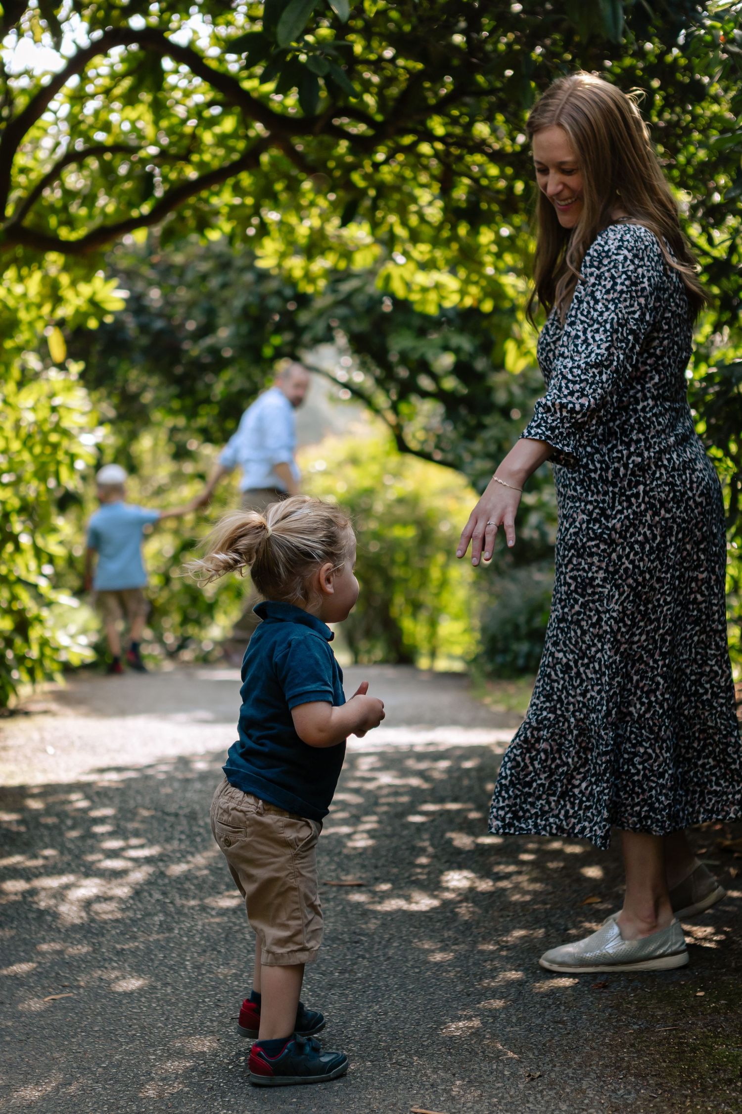 Toddler standing looking up at himother, mother smiling reaches out hand to him under arch in park, father and brother in the background
