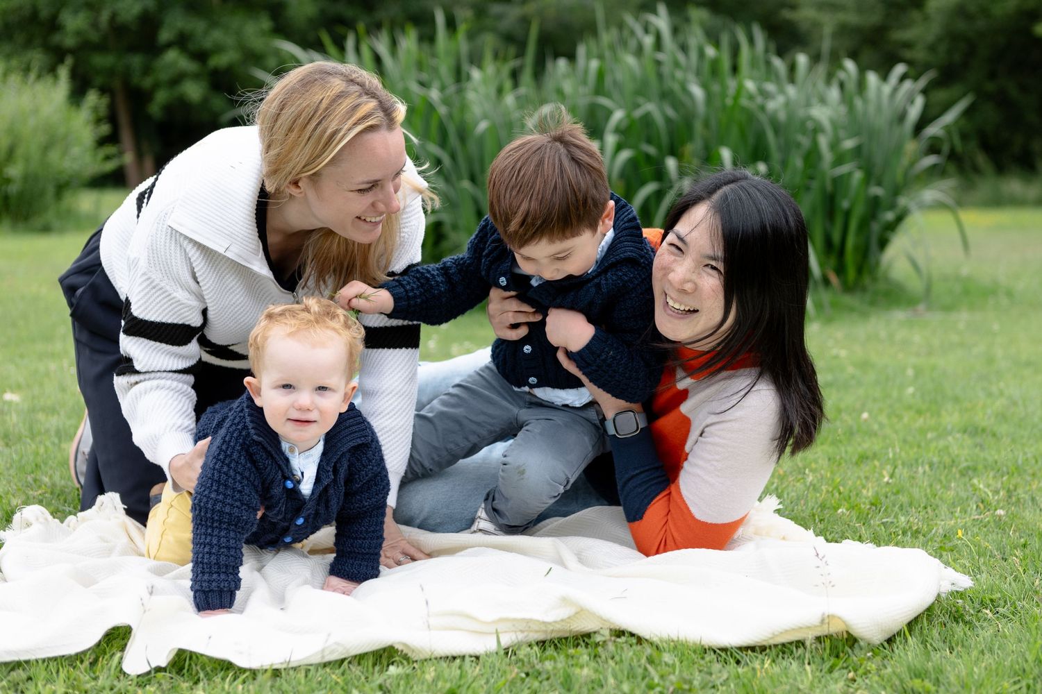 Family of four on blanket in park, with mothers holding and tickling their two toddler sons