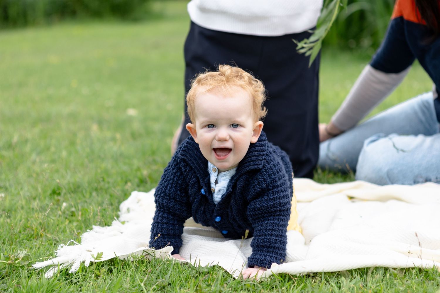 Baby boy on blanket in park crouching on all fours looking at camera with a big smile