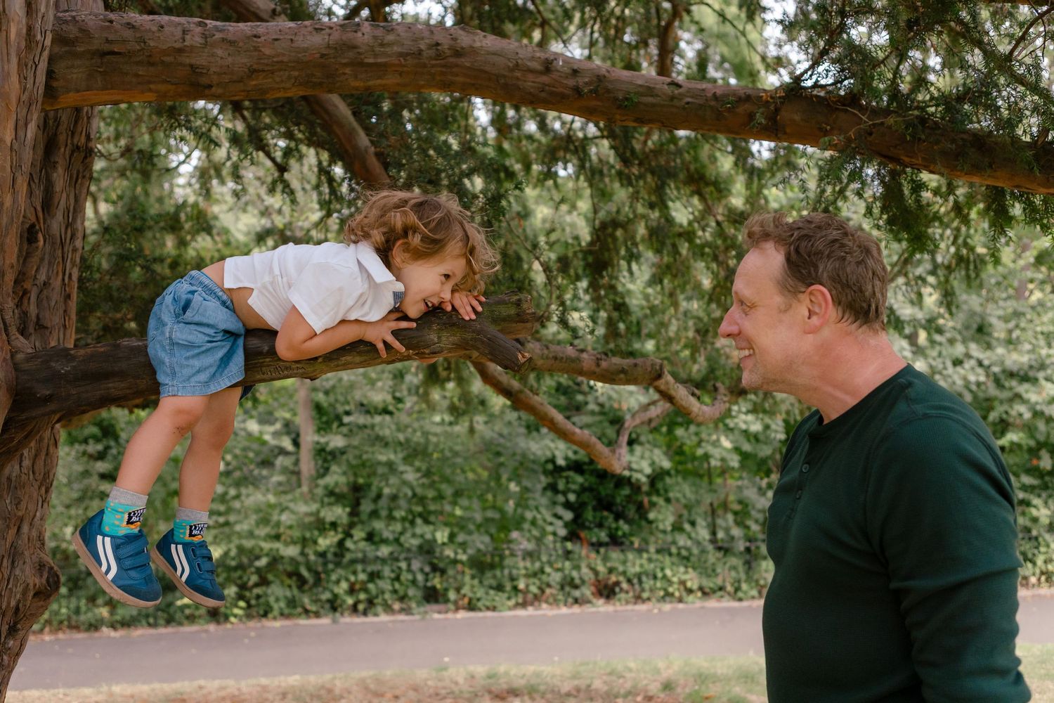 Toddler on a tree branch in park looking at his father and smiling, while father watches on and smiles back
