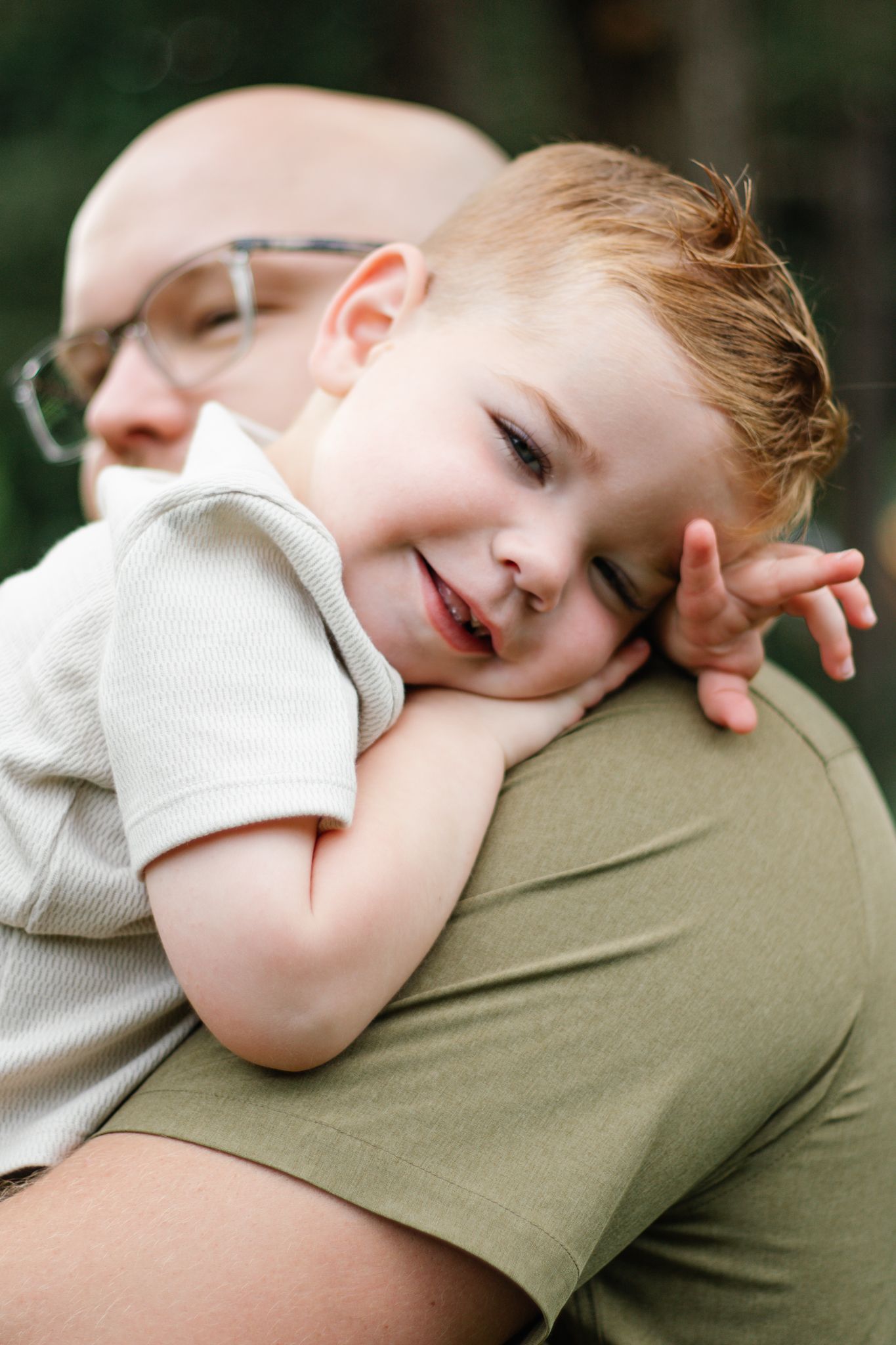 A tender moment captured as a child rests peacefully on a parent's shoulder in a green outdoor setting.