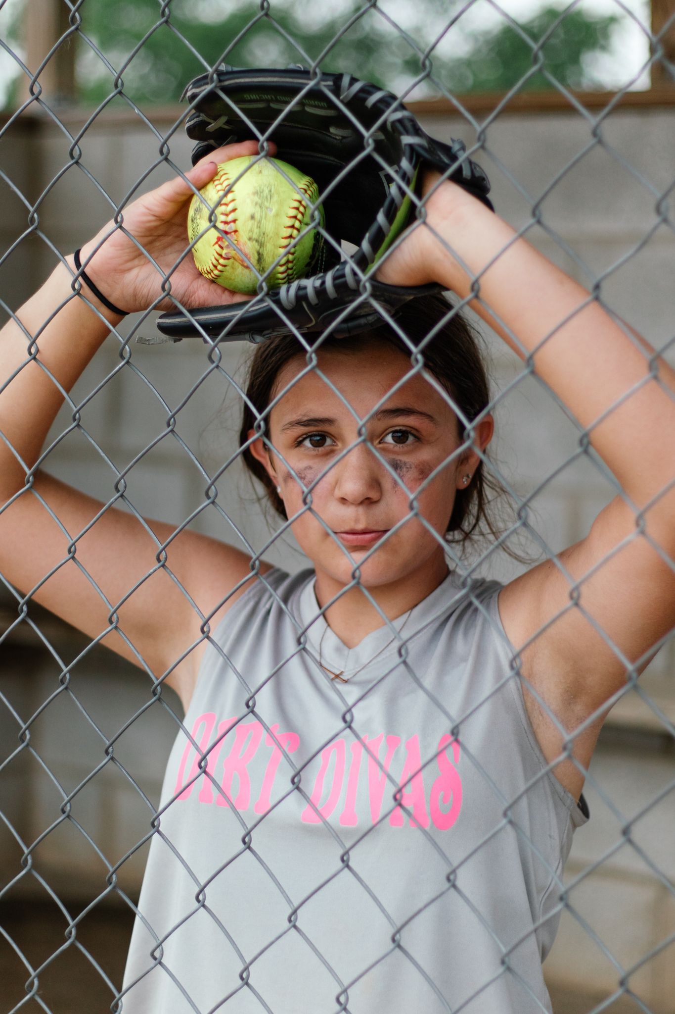 Children pose playfully against a chain link fence while holding softballs.
