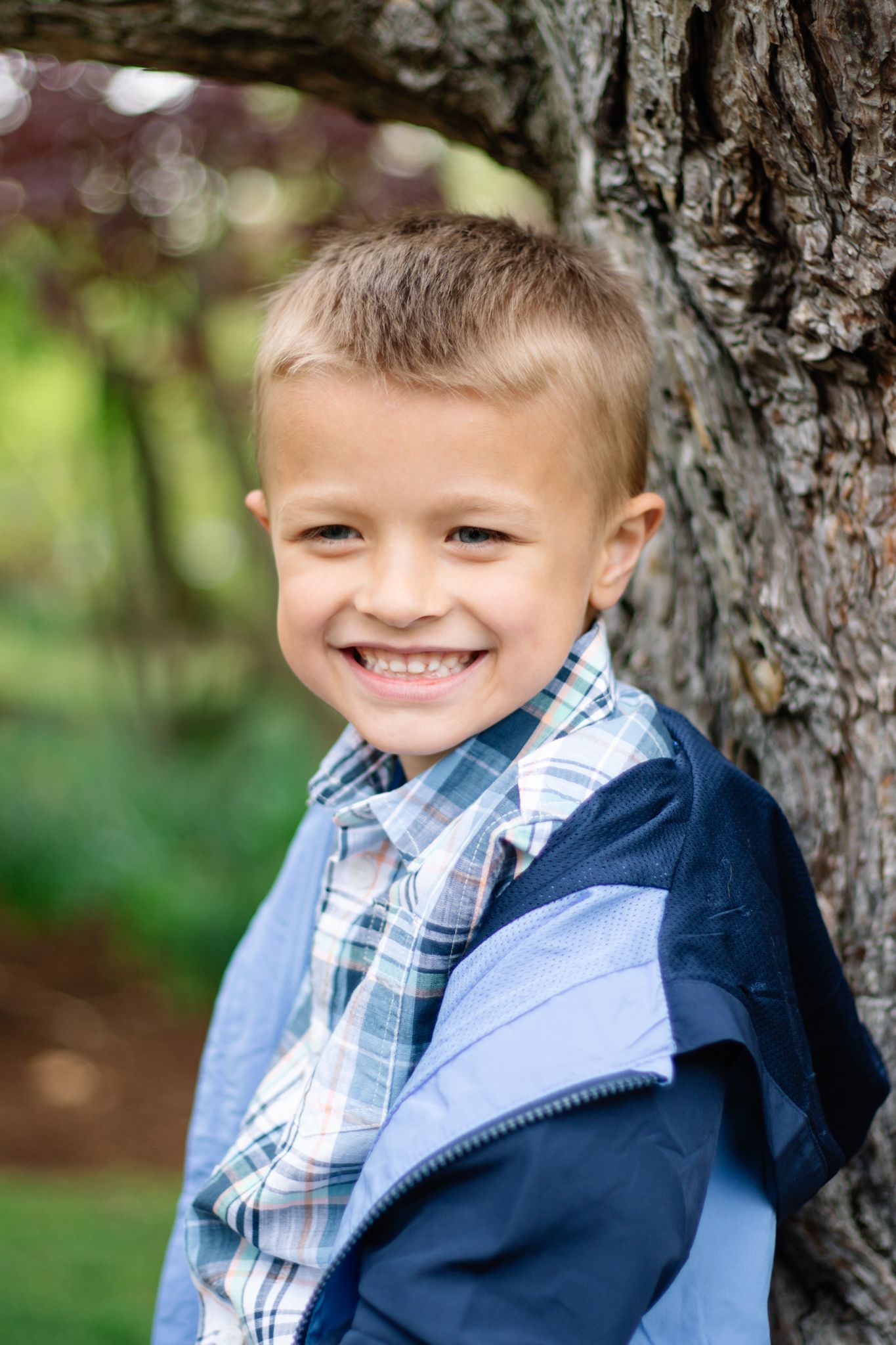 A child in a plaid shirt and navy jacket smiles while sitting near a tree trunk.