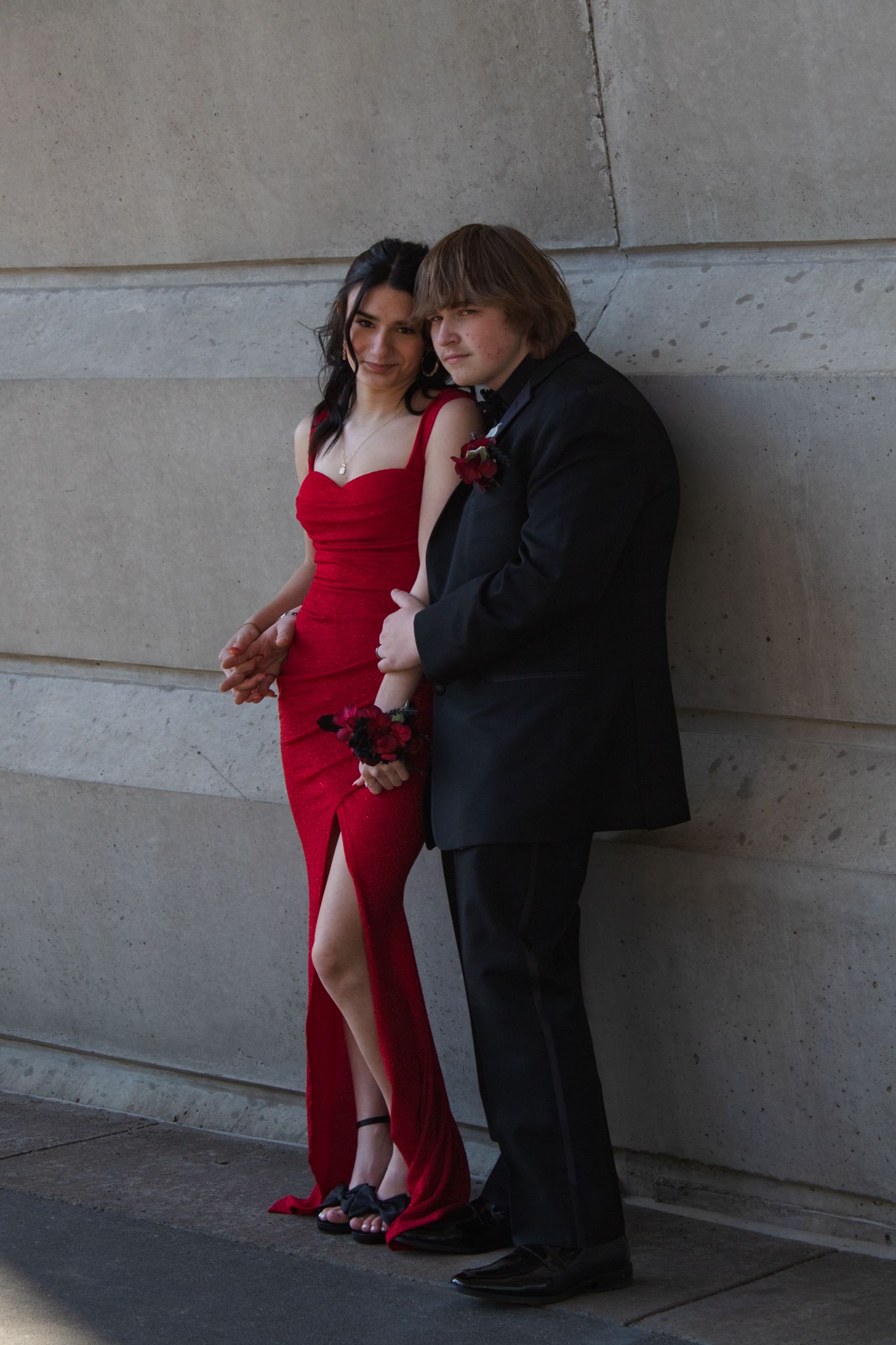 Formal couple poses against concrete wall, woman in red gown and man in black suit.