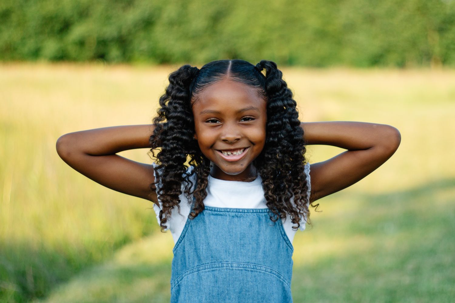 Young child in blue overalls plays joyfully in a sunny field with green grass and natural lighting.