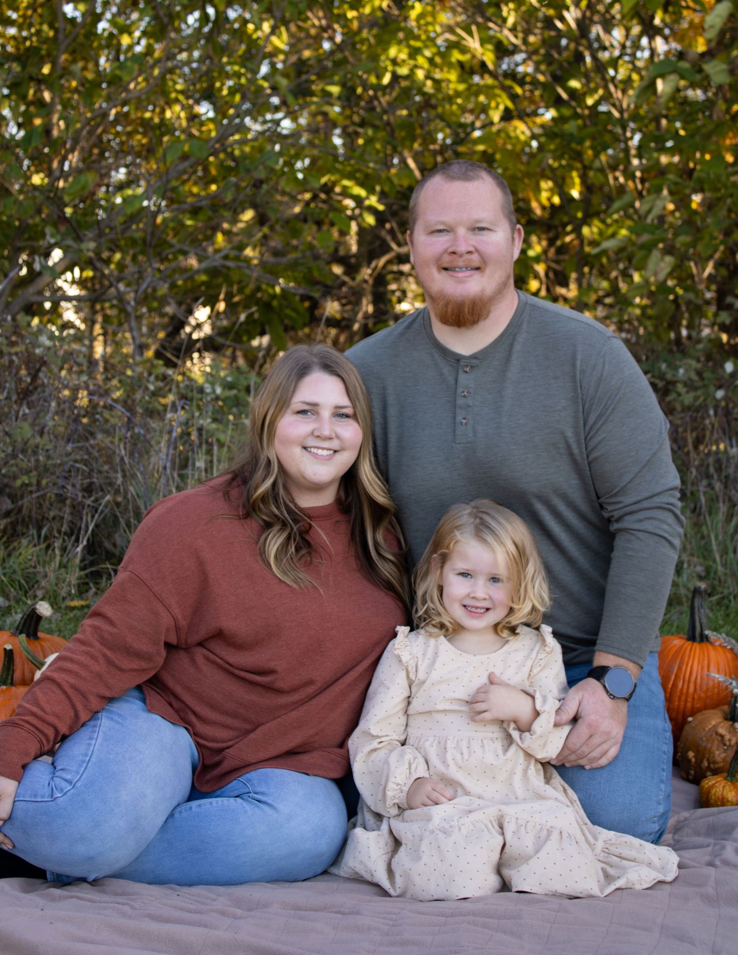 Family sits together outdoors in casual attire against a backdrop of fall trees and pumpkins.
