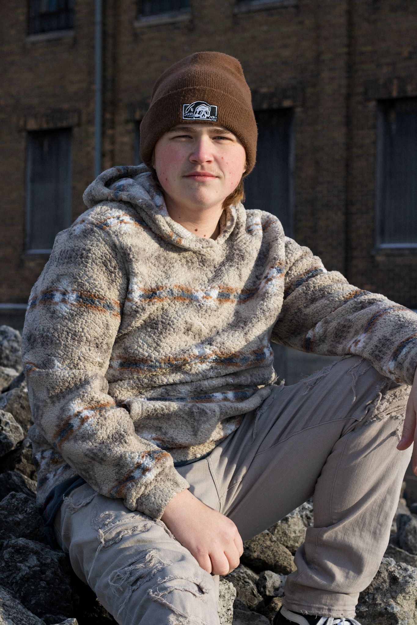 Person in brown beanie and fuzzy sweater sits against brick building in winter.