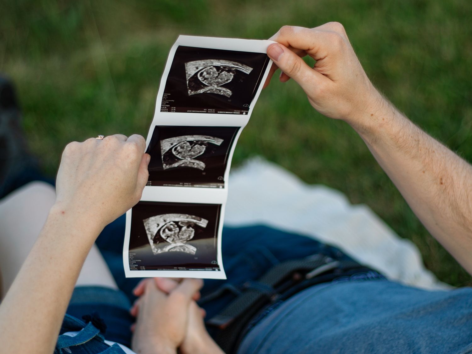 Couple lying in grass holding ultrasound photos in a time-lapse pregnancy announcement series.