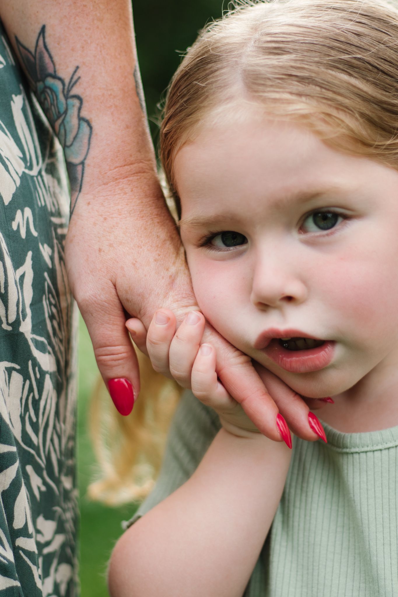 Child in sage green shirt smiles while playfully hiding behind adult hand.