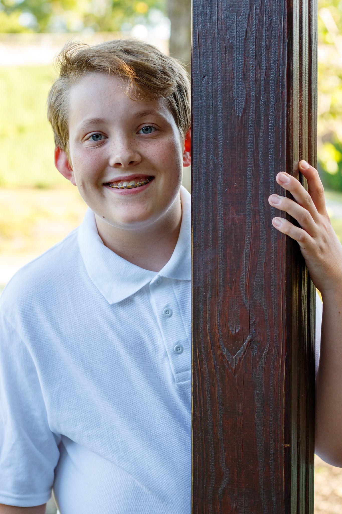 A person in a white polo shirt poses beside a dark wooden post outdoors.
