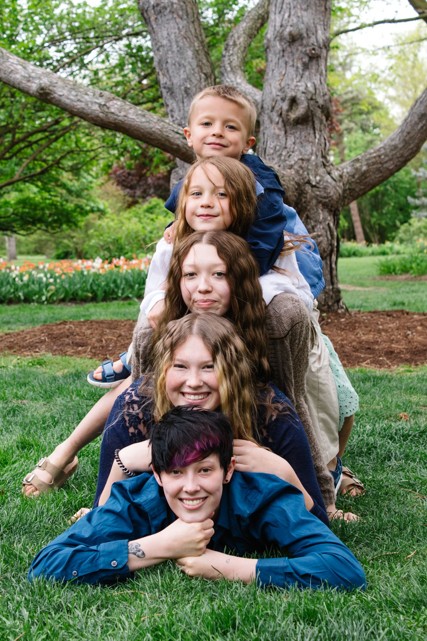Playful family members lying on grass under tree branches during outdoor photo session.