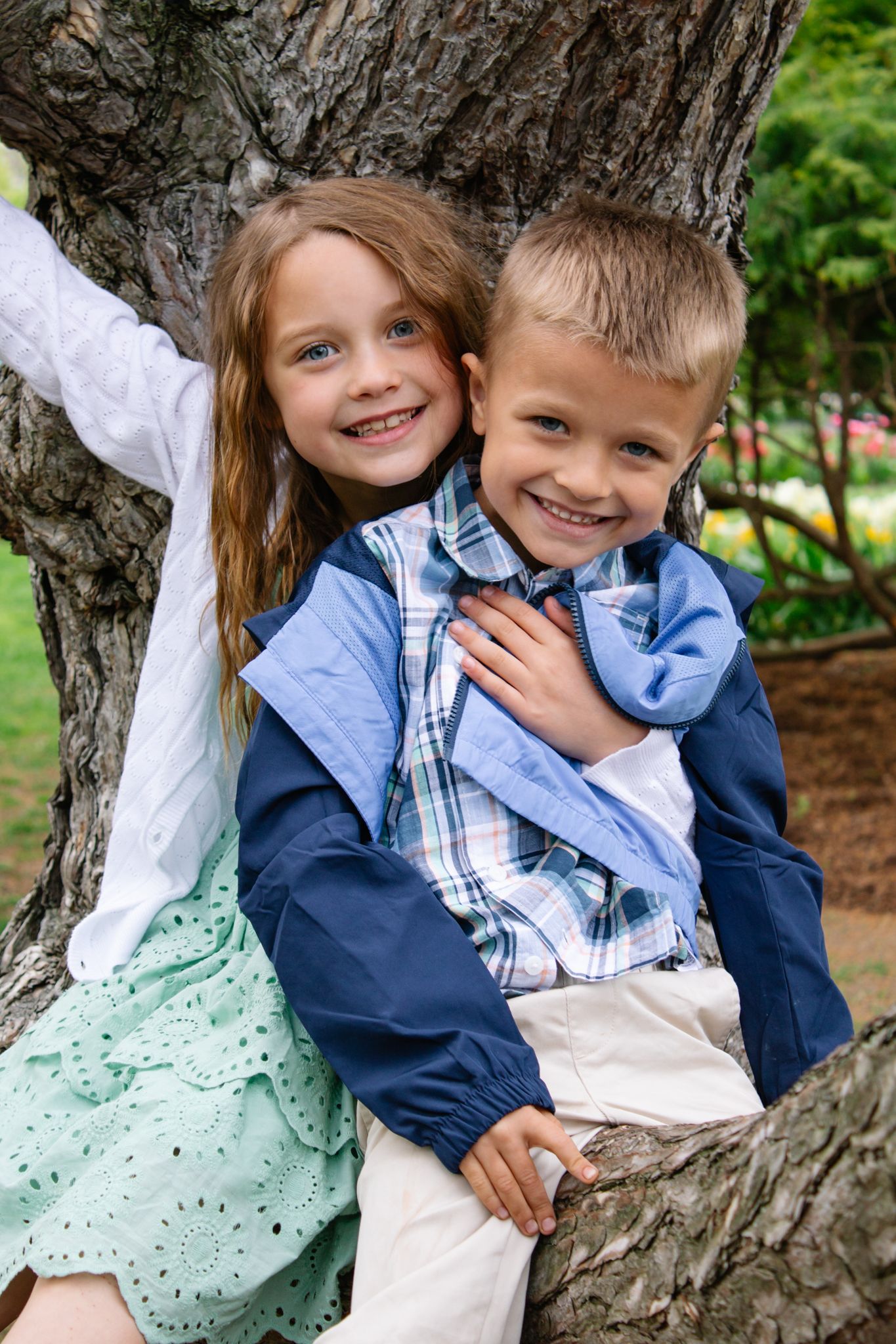 Siblings posing playfully by tree trunk in springtime garden setting.