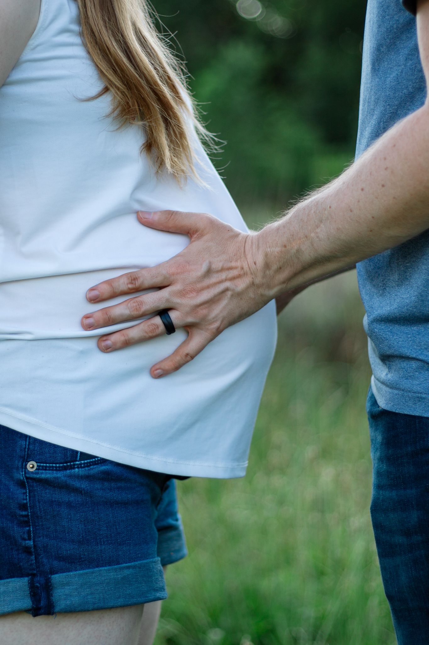 A couple holding hands over a pregnant belly in an outdoor setting.
