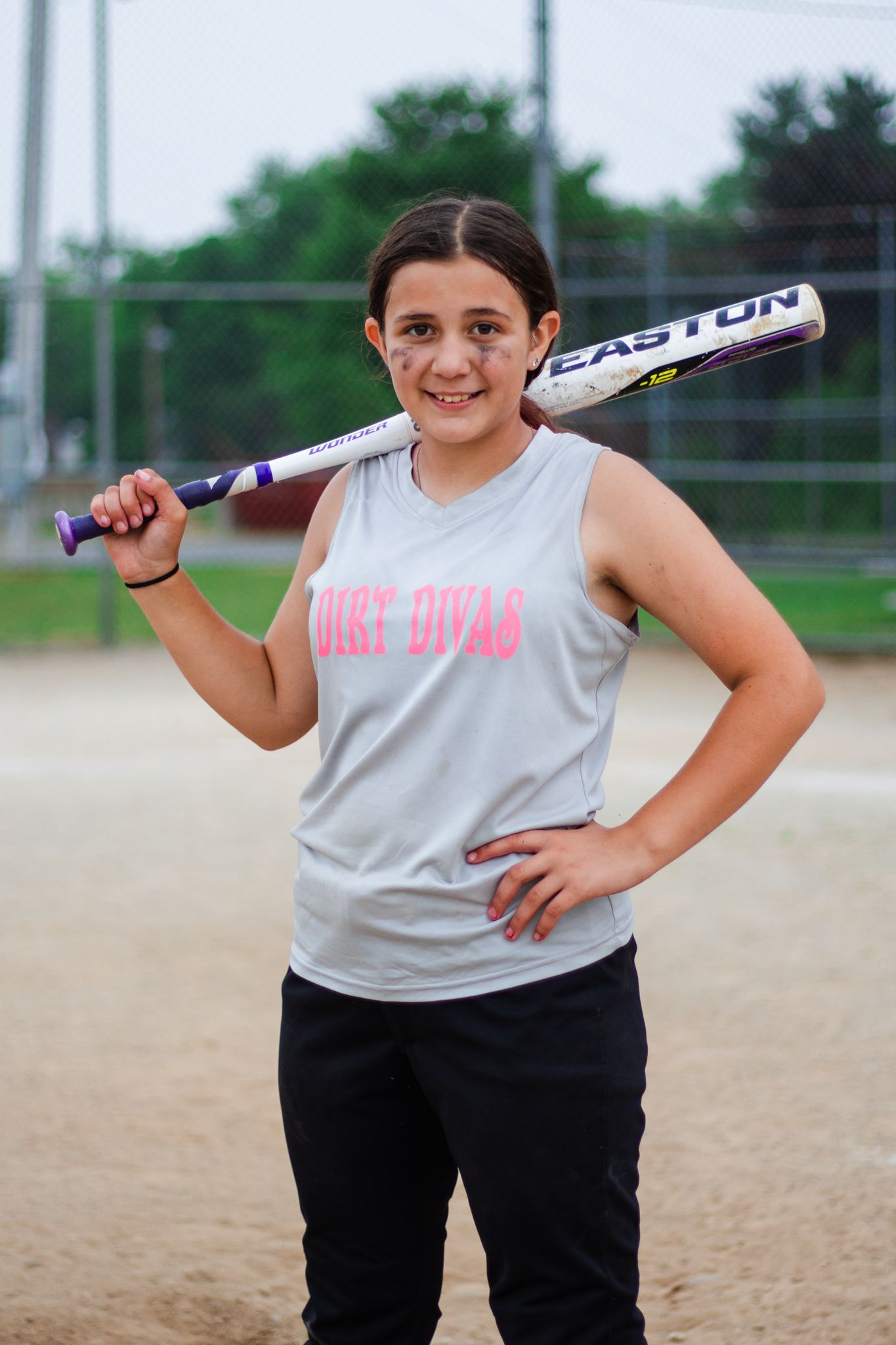 Softball player in gray shirt and black pants poses with bat on baseball diamond.