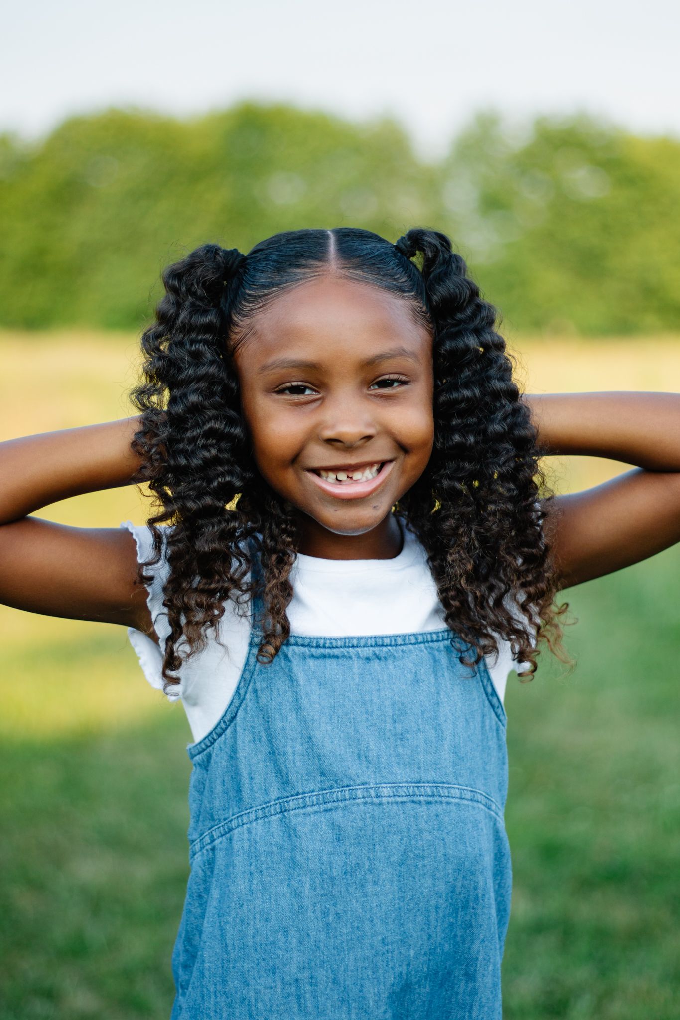 Child in denim dress posing playfully with hands behind head outdoors.