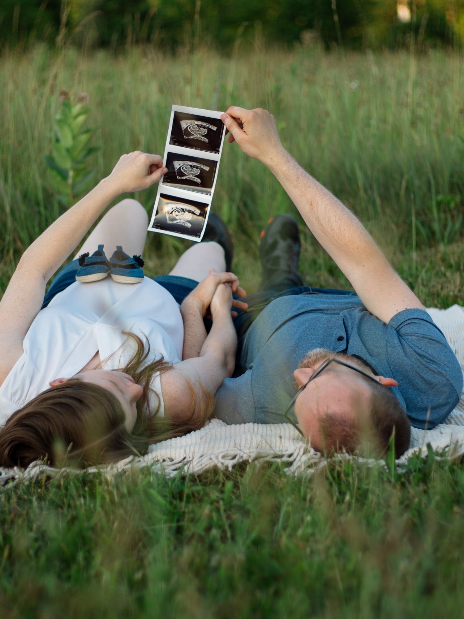Couple laying in grass holding ultrasound prints above them while relaxing outdoors.