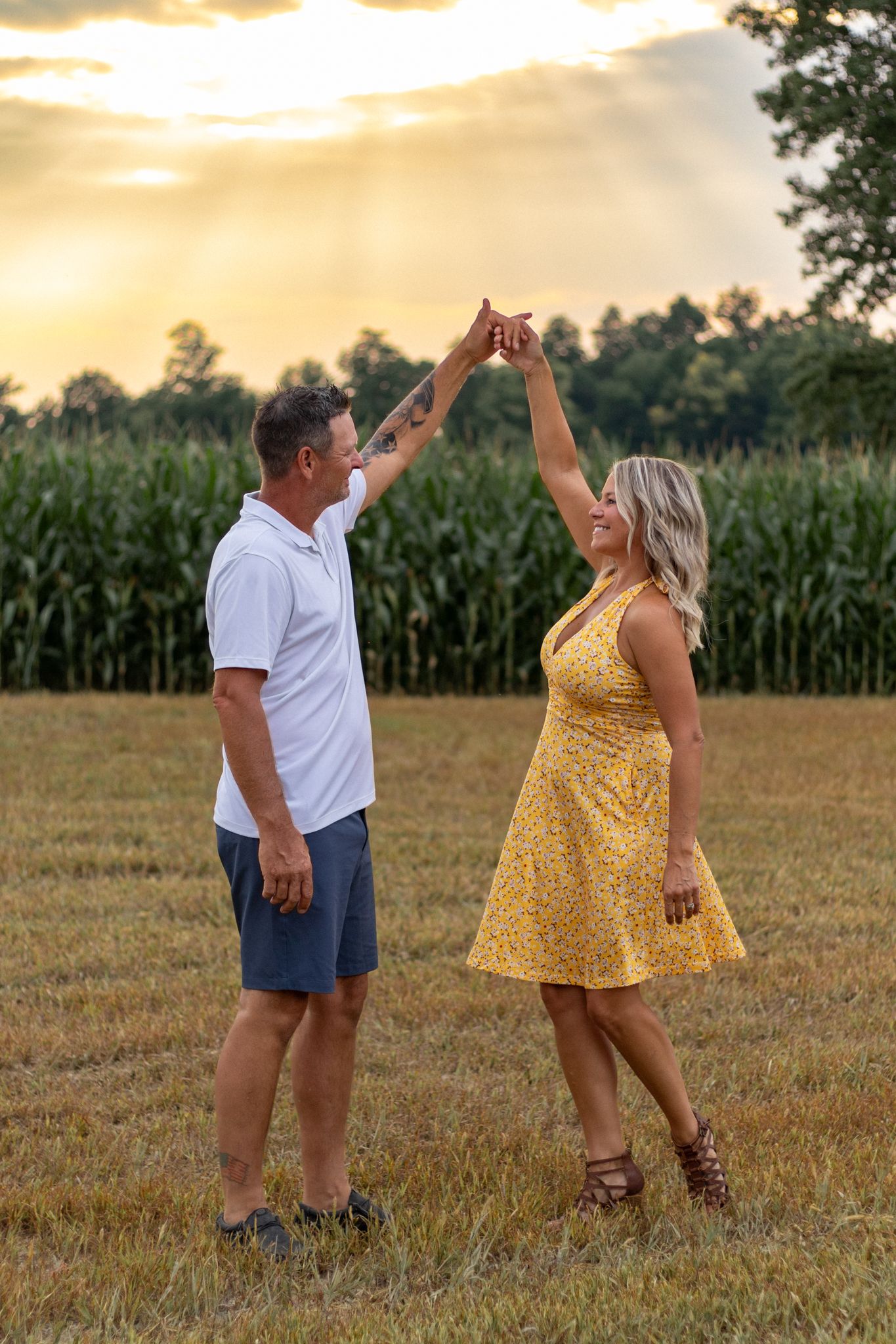 Couple dances playfully in a field at sunset with cornfields in the background.