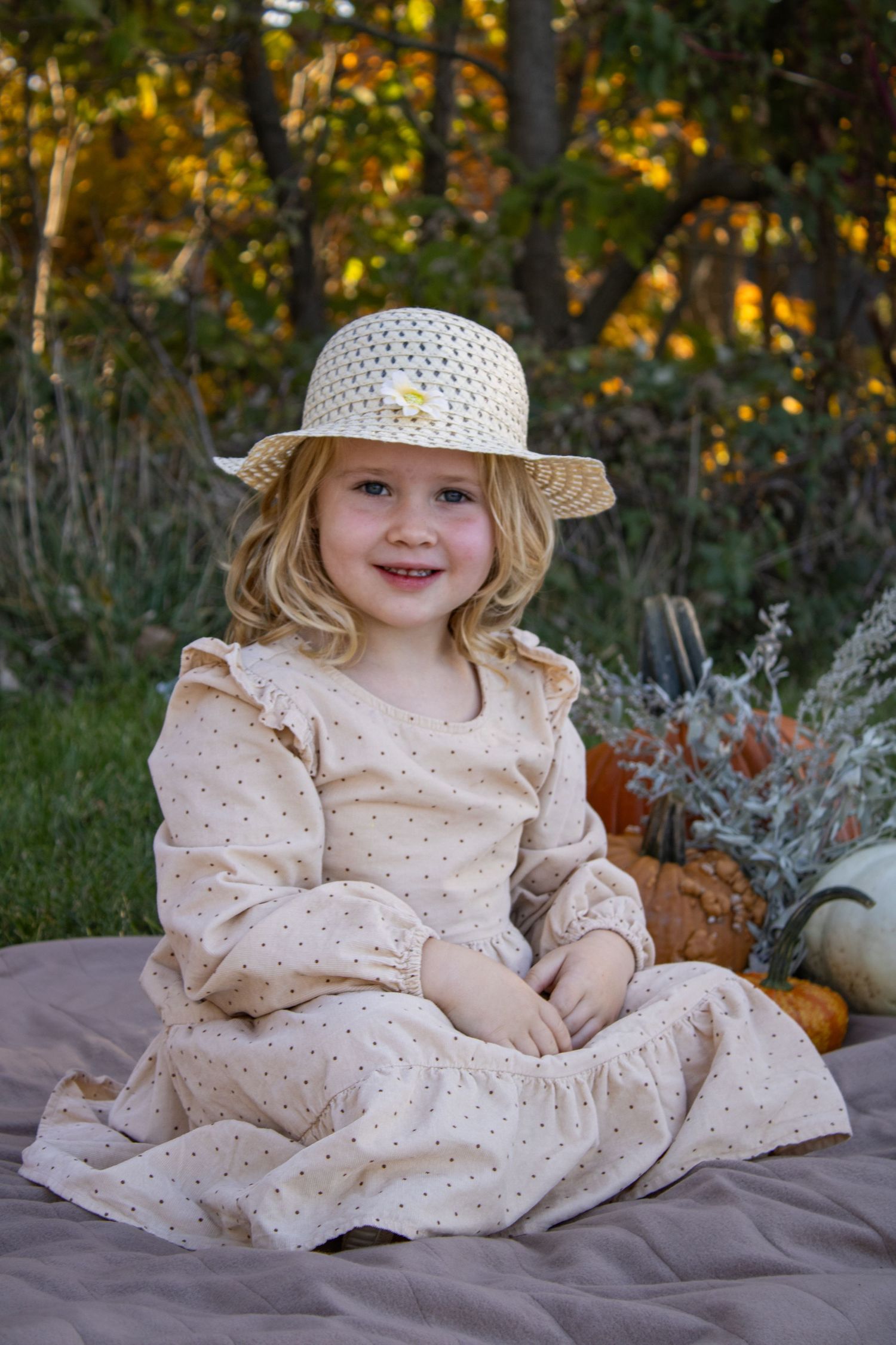 A person in a flowing cream dress and white sun hat poses among fall decorations.