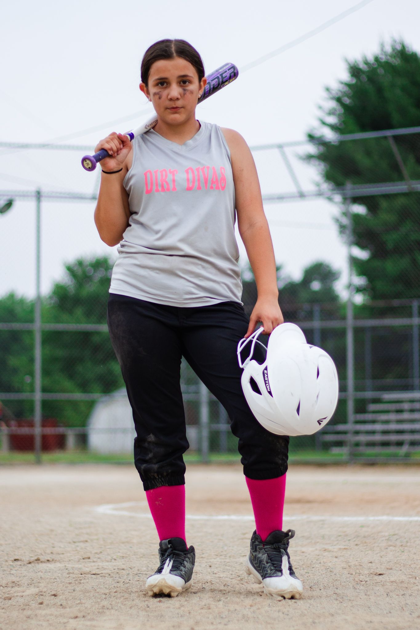 Softball player wearing pink socks and gray shirt holds bat and helmet on dusty field.